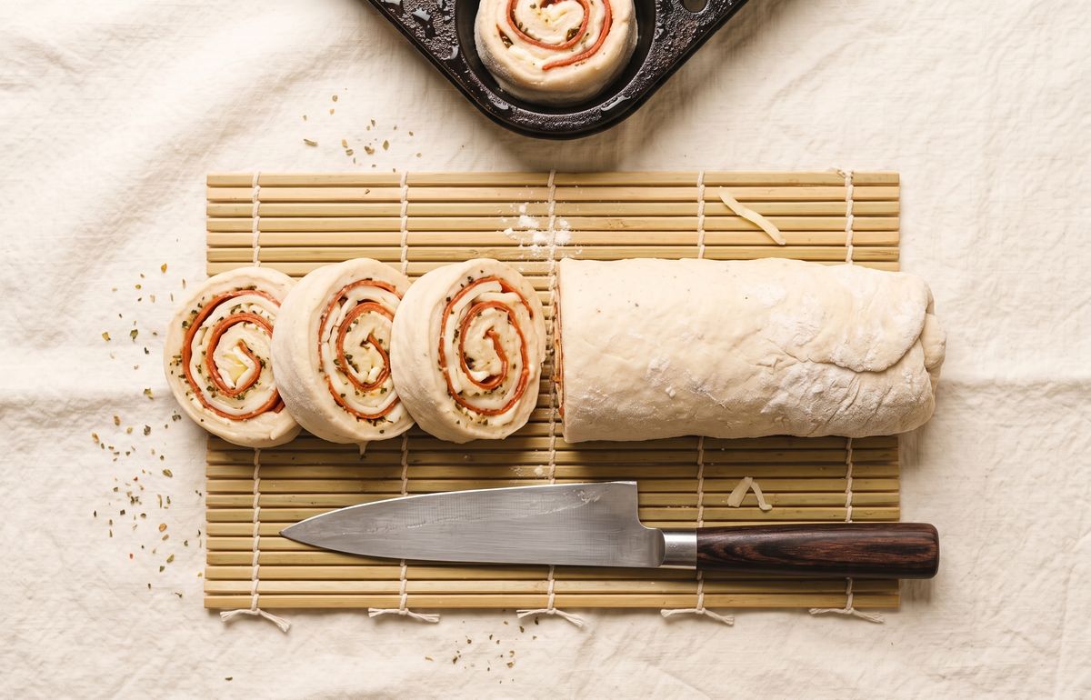 The rolled log sliced into rounds showing pepperoni and cheese spirals, with a chef's knife and a cast-iron pan