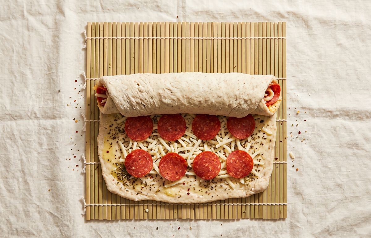 Sourdough dough brushed with butter and seasoning, topped with mozzarella and pepperoni, half rolled on a bamboo sushi mat